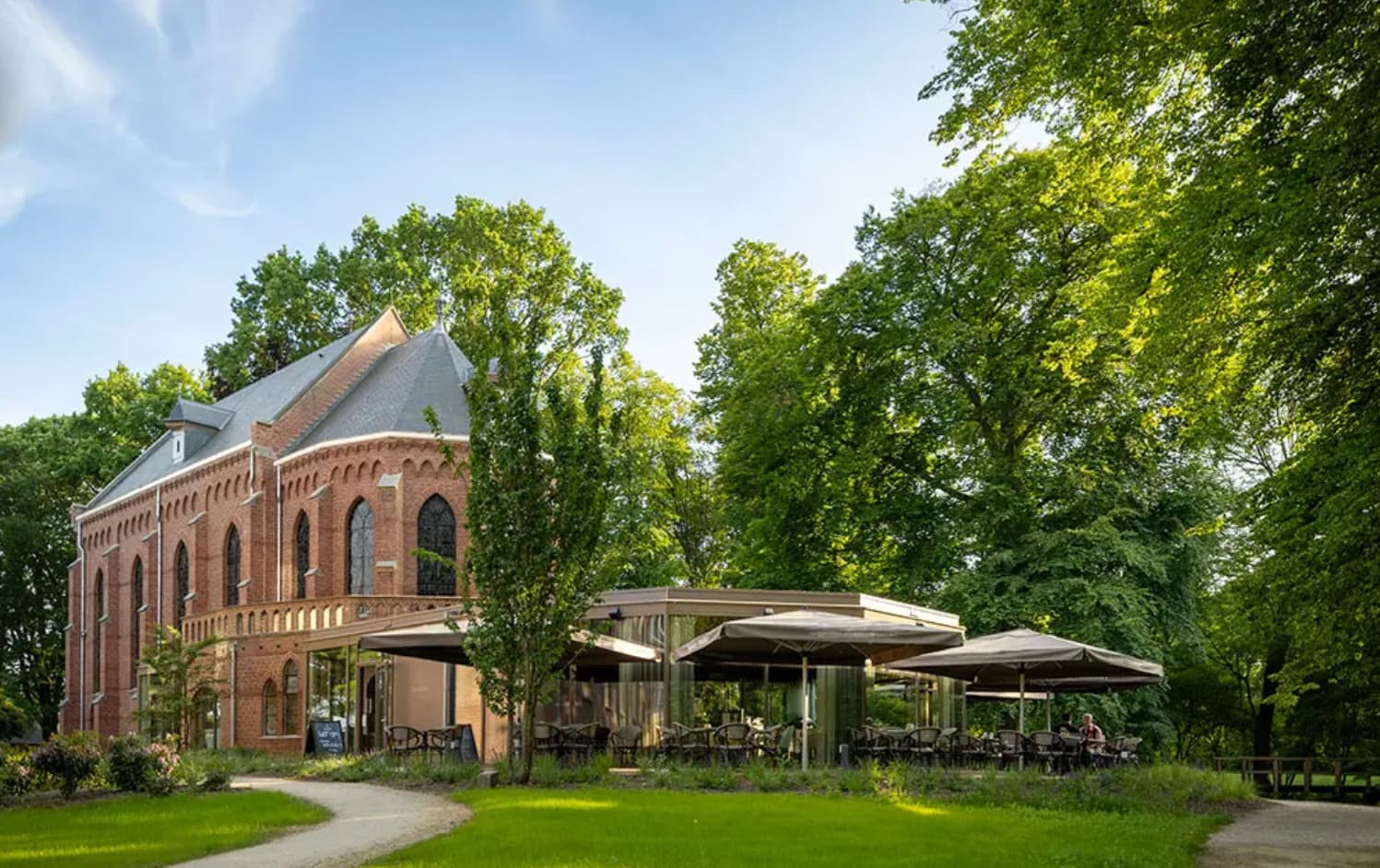Kapel Montfort van rode baksteen met gotische ramen tussen hoge bomen op Landgoed Bijstervelt, met het terras van de brasserie onder parasols ernaast, in de zon.