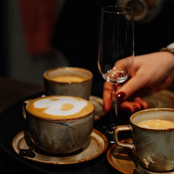 Een hand met rode nagellak houdt een leeg glas vast boven drie kopjes cappuccino in grijs aardewerk.